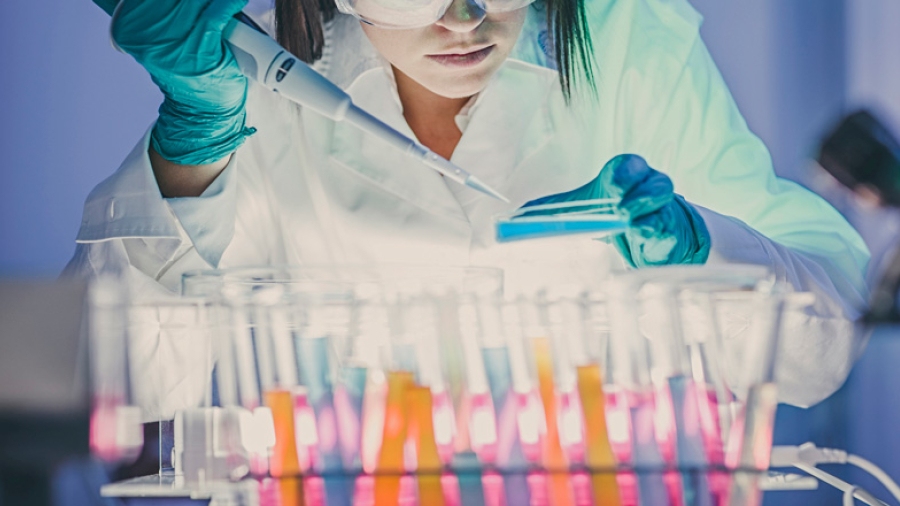 A laboratory worker holding a pipette and a Petri dish, behind a row of test tubes